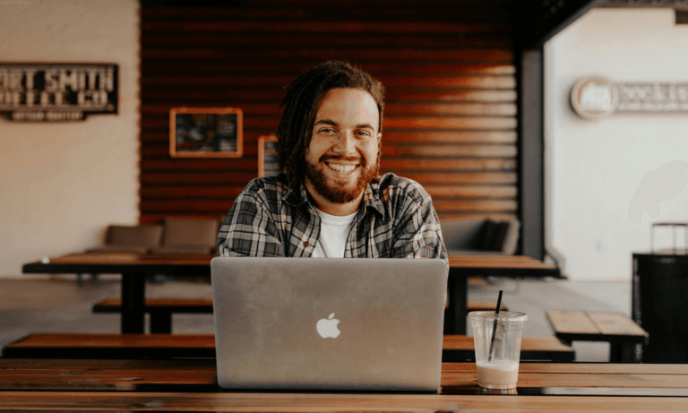 A man in a cafe with his laptop, smiling at the camera